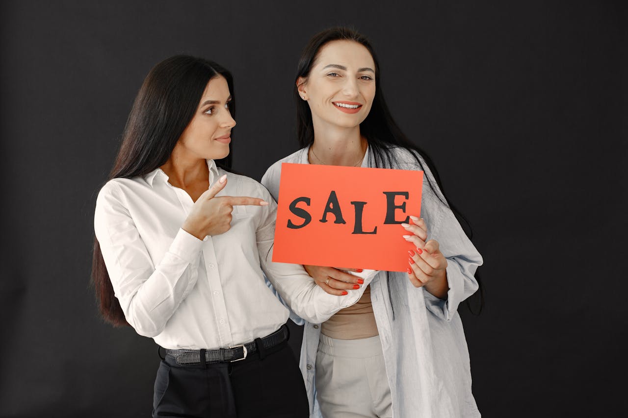 Two women with long hair smiling and holding a sale sign against a black background.